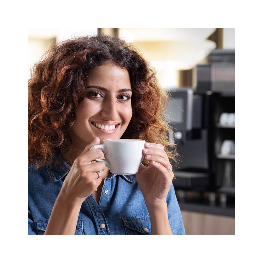 A person with curly hair smiles while holding a coffee cup near their mouth. They are wearing a denim shirt and sitting in a cafe setting with blurred coffee machines in the background