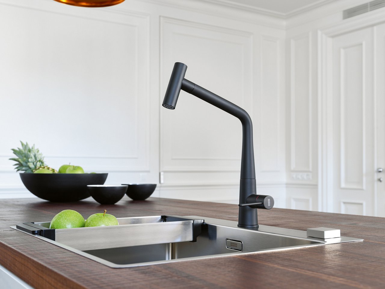 Franke Icon Matte Black pull out kitchen faucet in paired with a stainless steel sink in a contemporary kitchen feature dark wood counters. Sink has a colander with apples 