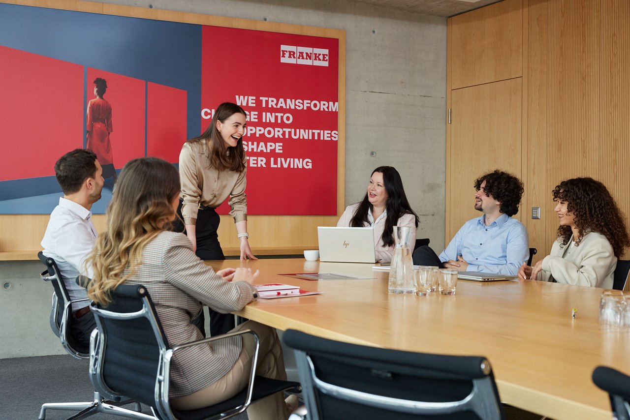 Group meeting in a modern conference room with a large Franke wall poster.