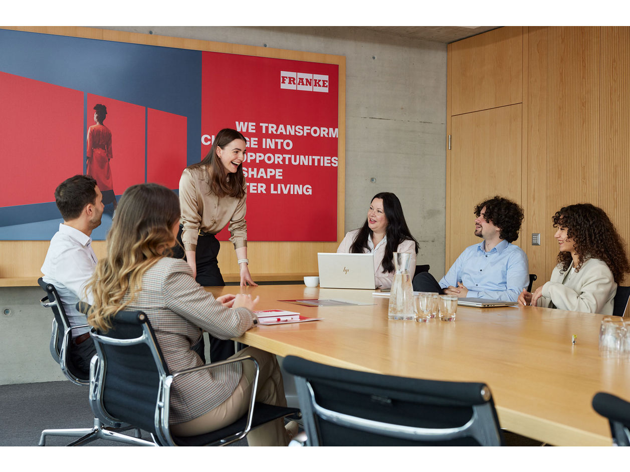 Group meeting in a modern conference room with a large Franke wall poster.