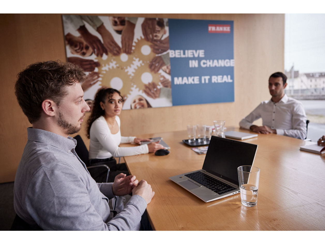 Four people seated at a conference table in a meeting room with a laptop and glasses of water. A poster on the wall reads "Believe in Change, Make it Real