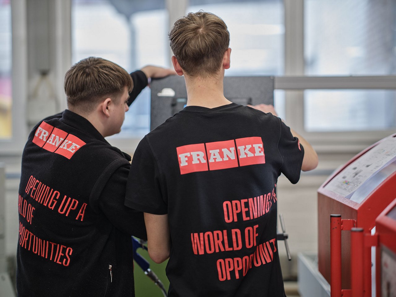 Two young men in a workshop, wearing black clothing with red "FRANKE" branding, facing a machine