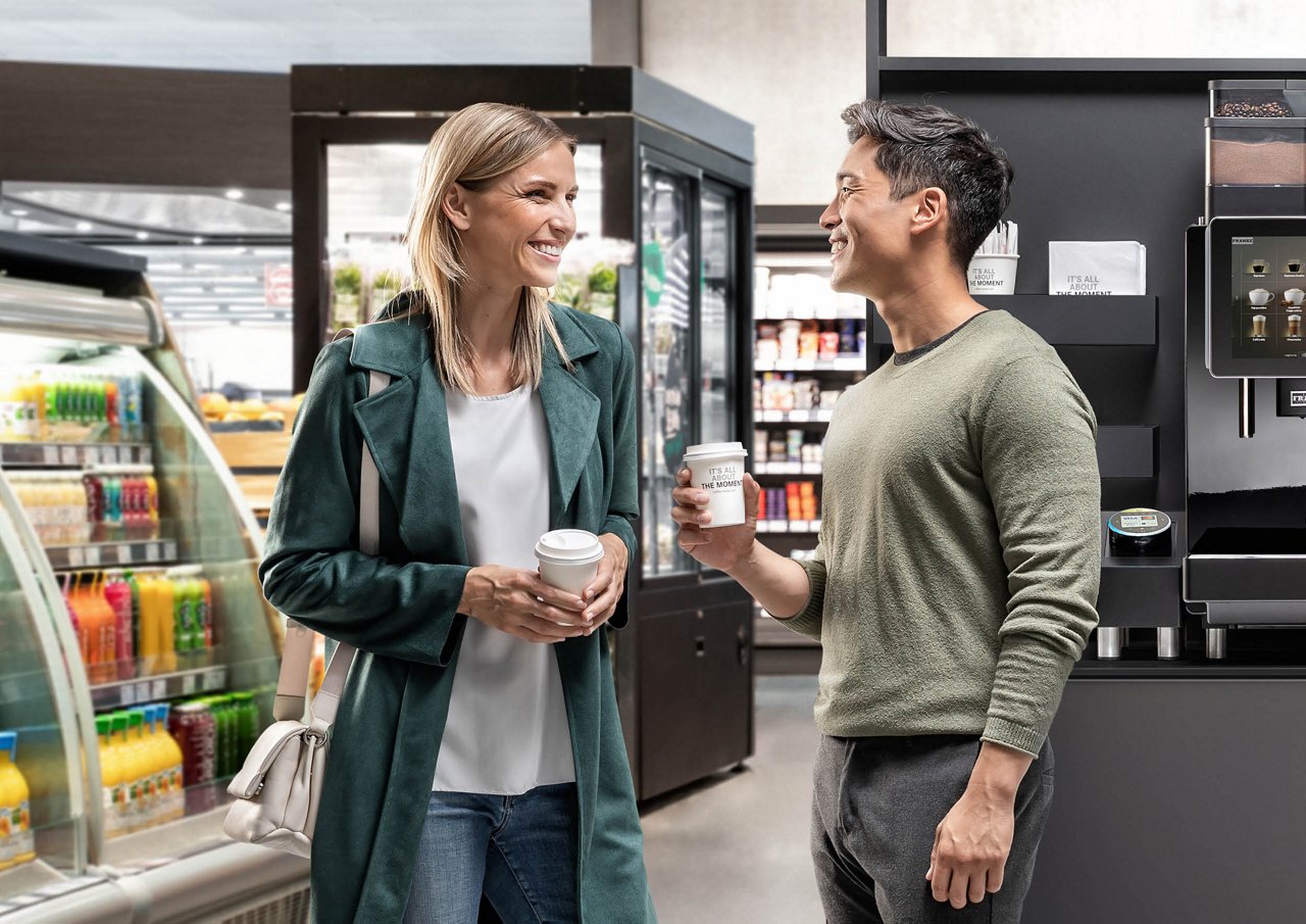 Franke Coffee Systems, man and woman holding cups for coffee to go in convenience store, fully automatic coffee machine Franke in the background