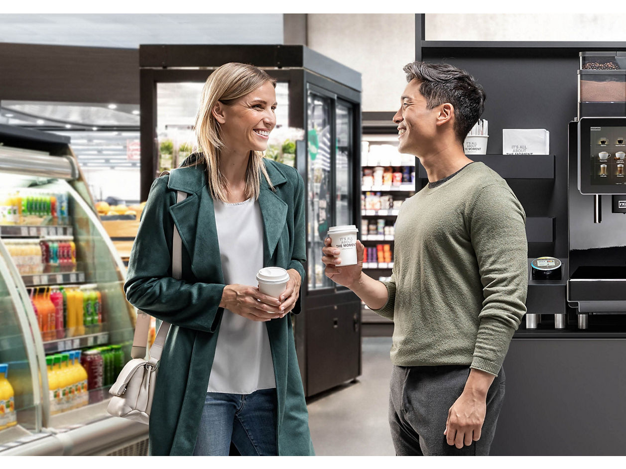 Franke Coffee Systems, man and woman holding cups for coffee to go in convenience store, fully automatic coffee machine Franke in the background