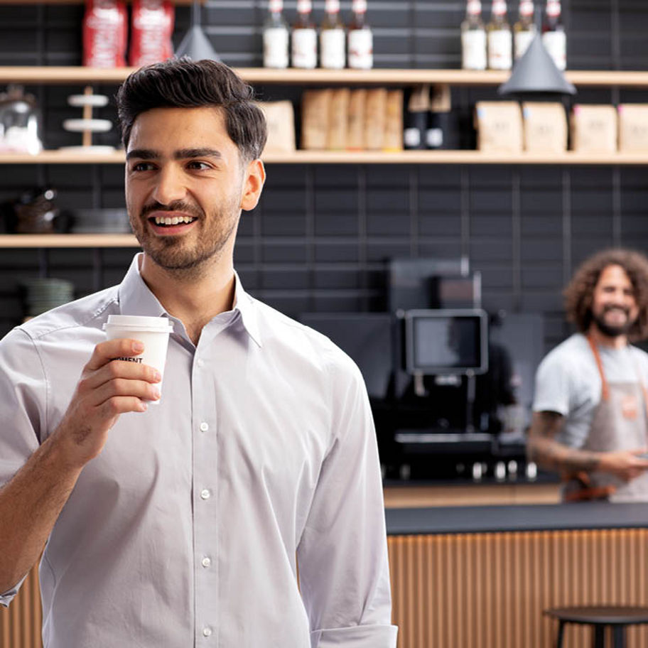 Franke Coffee Systems, man in coffee shop holding a coffee to go cup, counter and waiter near fully automatic coffee machine Franke in the background
