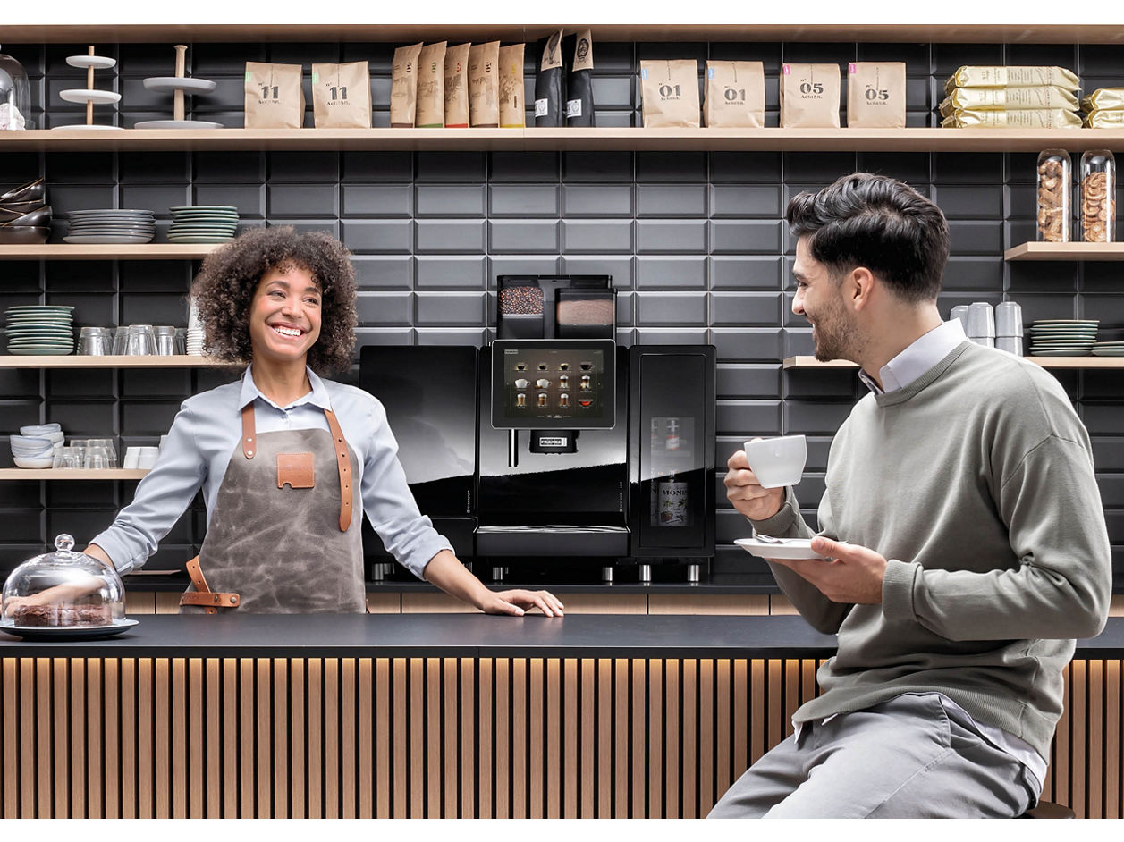 Franke Coffee Systems, man sitting at the counter of a coffee shop drinking coffee served by woman shop assistant, fully automatic coffee machine Franke 