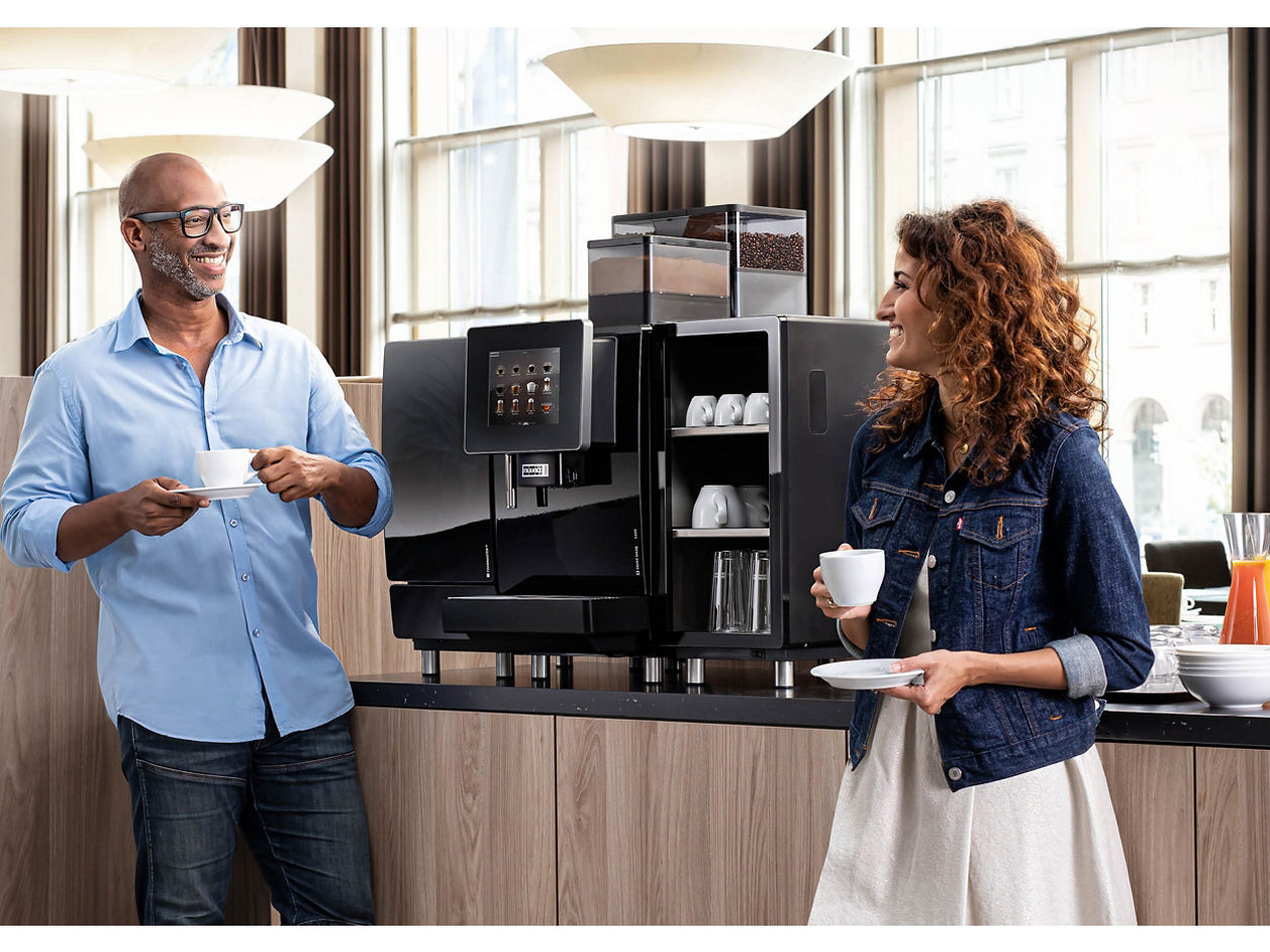 Franke Coffee Systems, man and woman holding cups in hotel breakfast area, in front of a fully automatic coffee machine Franke A600