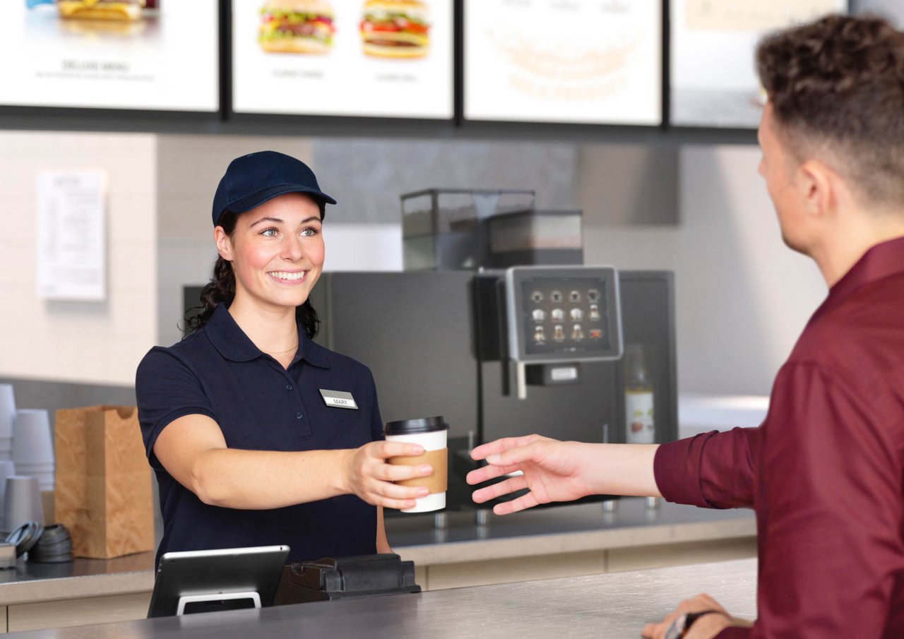 Franke Coffee Systems, man taking a coffee to go in quick service restaurant, served by waitress, fully automatic coffee machine Franke in the background