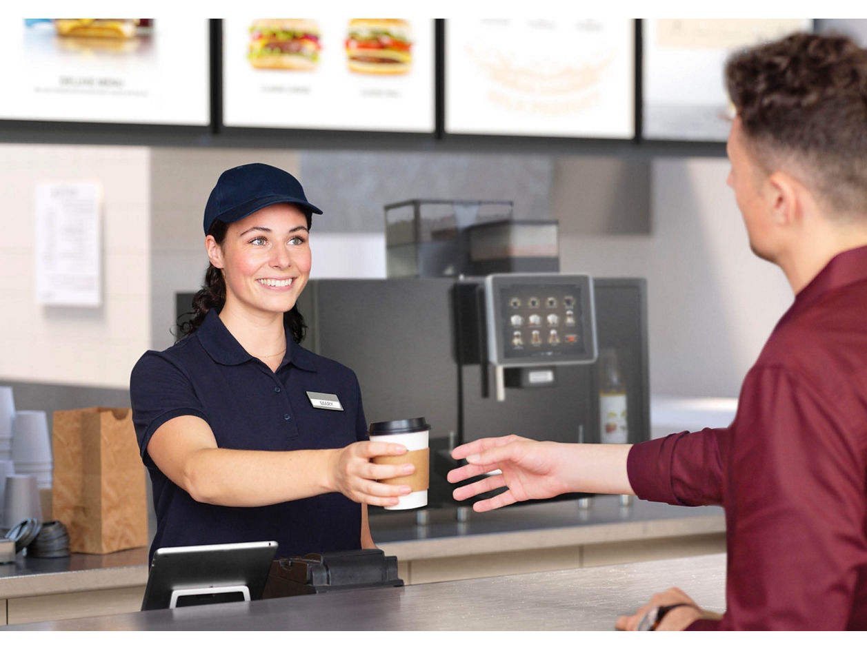 Franke Coffee Systems, man taking a coffee to go in quick service restaurant, served by waitress, fully automatic coffee machine Franke in the background