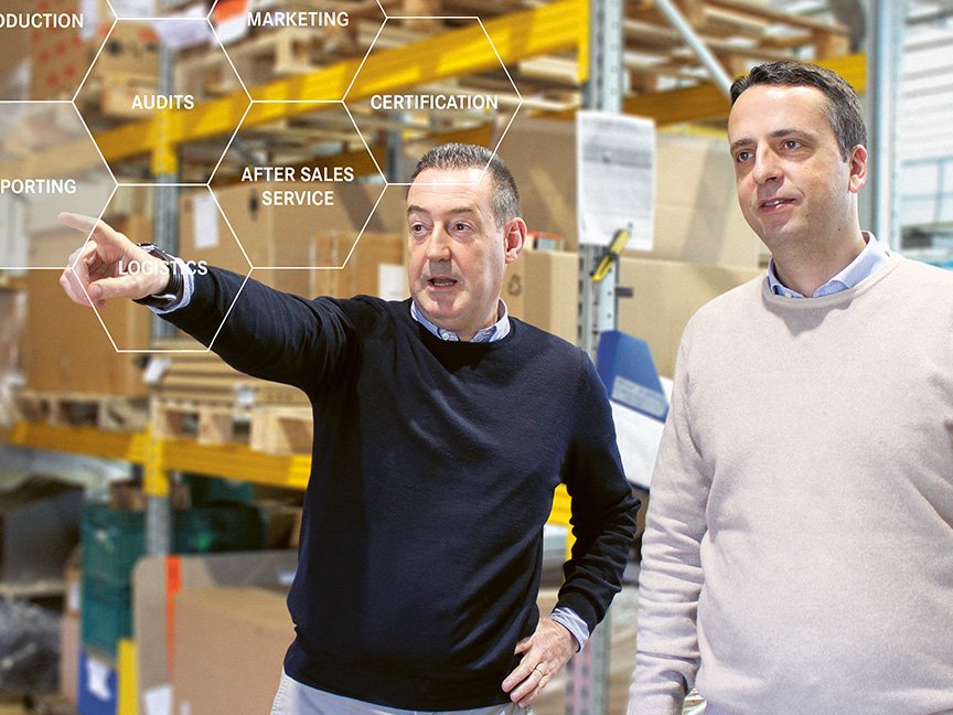 Two men in a warehouse observe something. One points ahead, while the other looks in the same direction. Overlay text includes terms like AUDITS and MARKETING. Shelves and industrial equipment are visible in the background