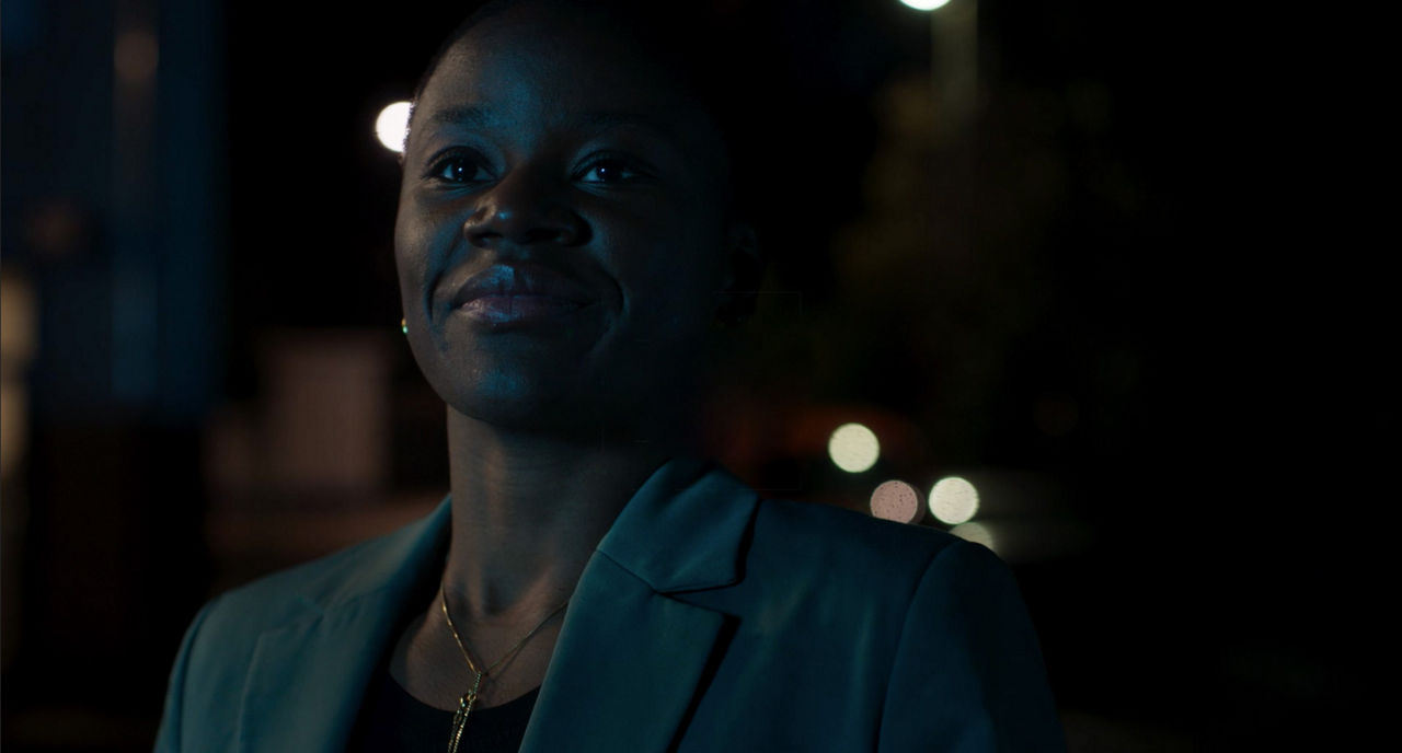 Person smiling softly in a dimly lit setting with blurred lights in the background