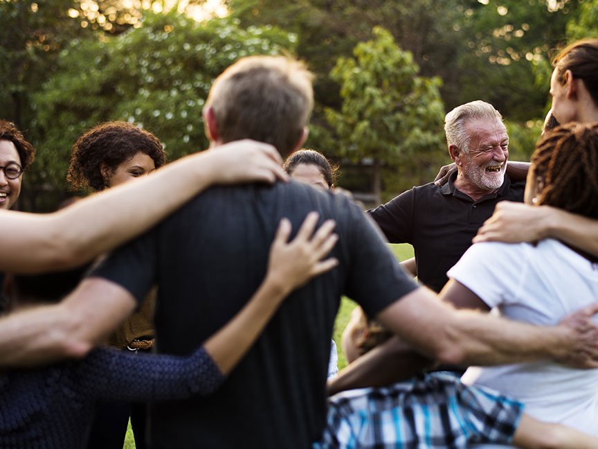 A diverse group of people stand in a circle outdoors, arms around each other’s shoulders, smiling and laughing. They appear to be enjoying a warm, sunny day in a park setting with trees in the background