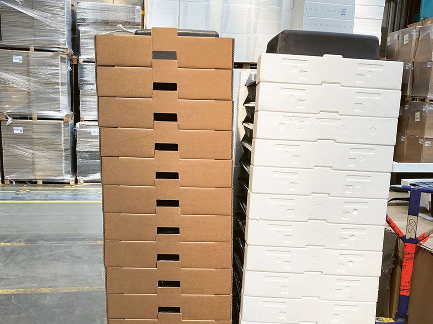 A stack of cardboard boxes is on the left, and a stack of white Styrofoam boxes is on the right. Both are on a warehouse floor with surrounded by wrapped pallets. A red forklift is partially visible at the bottom right