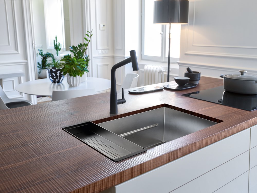 Franke Icon Matte Black pull out kitchen faucet in paired with a stainless steel sink in a contemporary kitchen feature dark wood counters. Sink has a colander with apples 