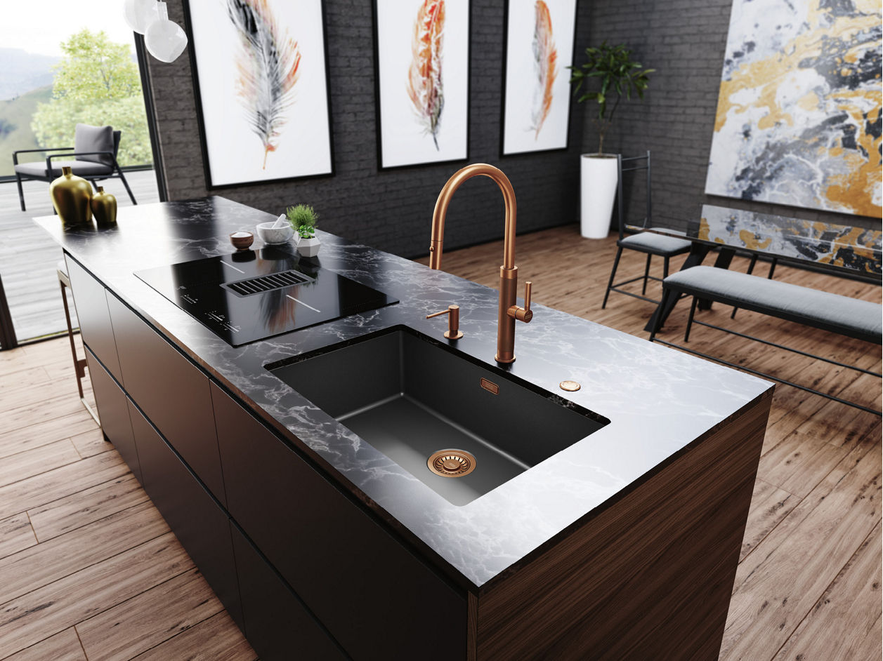 Nowoczesna kuchnia z dużą wyspą z czarnym blatem, w którym znajduje się zintegrowany zlewozmywak i miedziana bateria oraz płyta indukcyjna; w tle widoczne są trzy obrazy z motywem piór, abstrakcyjna grafika, ławka z poduszkami, drewniana podłoga i duże okna wpuszczające naturalne światło.