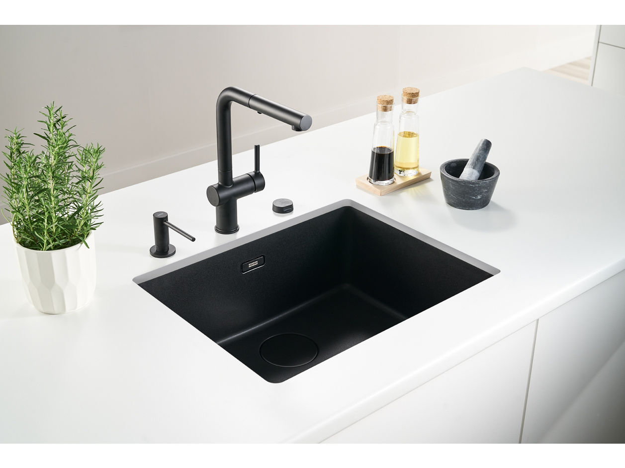 Czarny zlewozmywak granitowy wpuszczany w blat z jasnego kamienia, z czarną baterią kuchenną oraz tacą z oliwą i przyprawami ustawioną obok