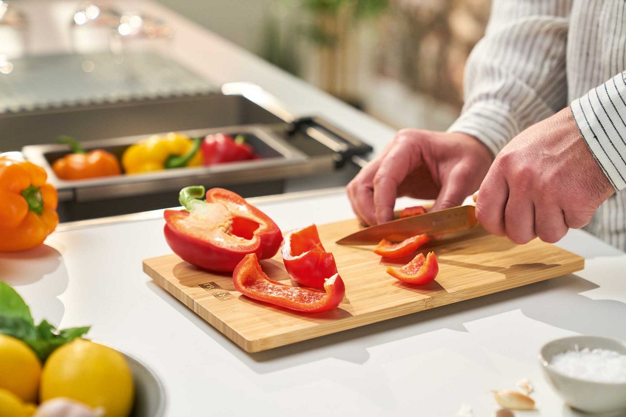 Red peppers being sliced on a bamboo cutting board next to a kitchen sink 