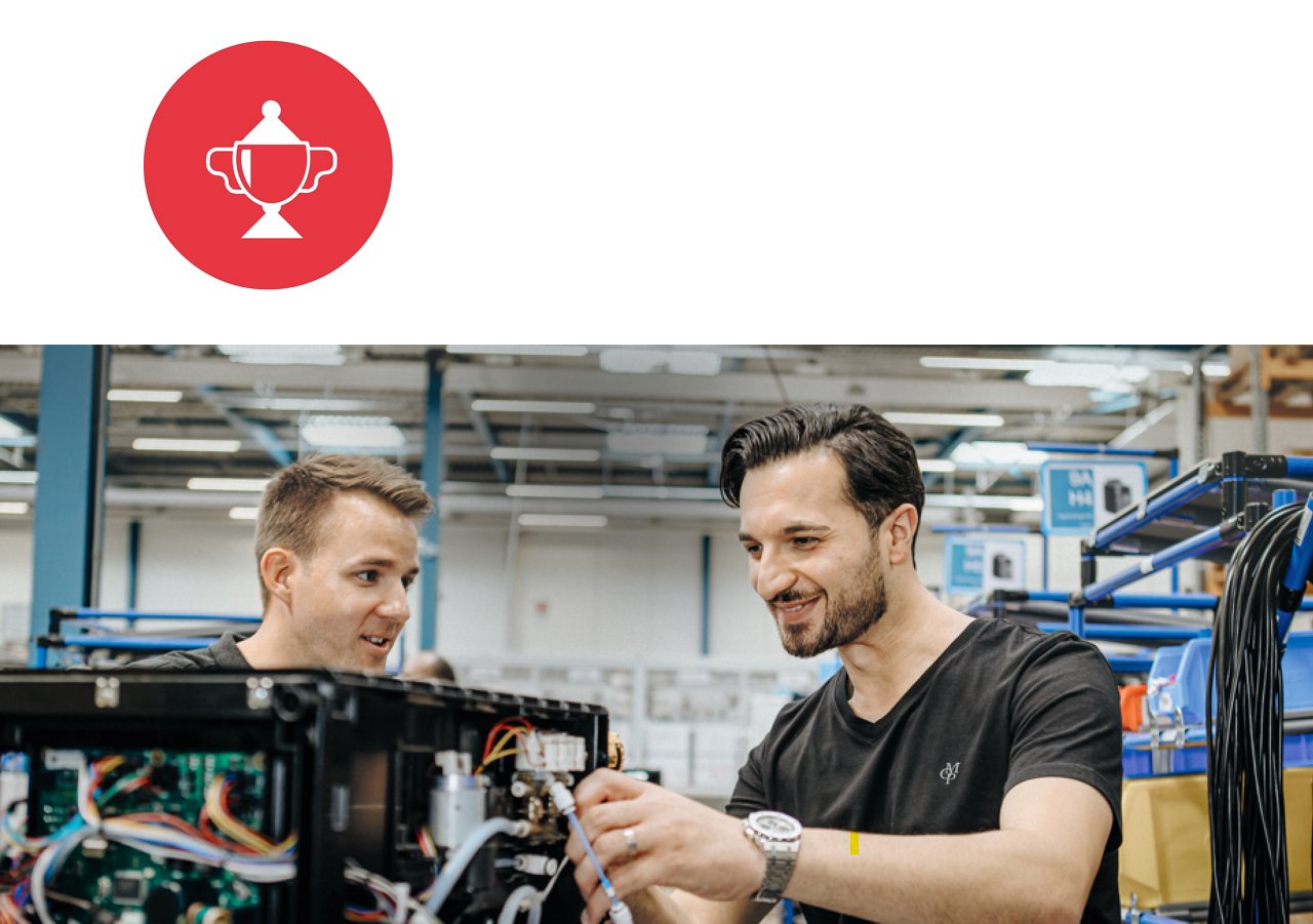 Two men working on electronic equipment in an industrial setting, with a red circle and white trophy icon above them.