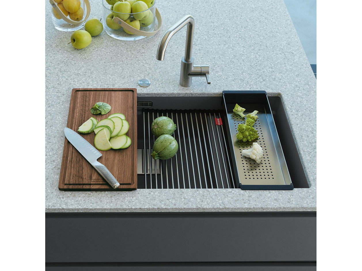 Nowoczesna strefa zmywania Franke z elegancką baterią ze stali nierdzewnej. Na blacie znajduje się drewniana deska do krojenia z zielonymi jabłkami i nożem, suszarka z całymi jabłkami oraz czarny durszlak z sałatą i czosnkiem. W tle widoczna miska z owocami na jasnym blacie.