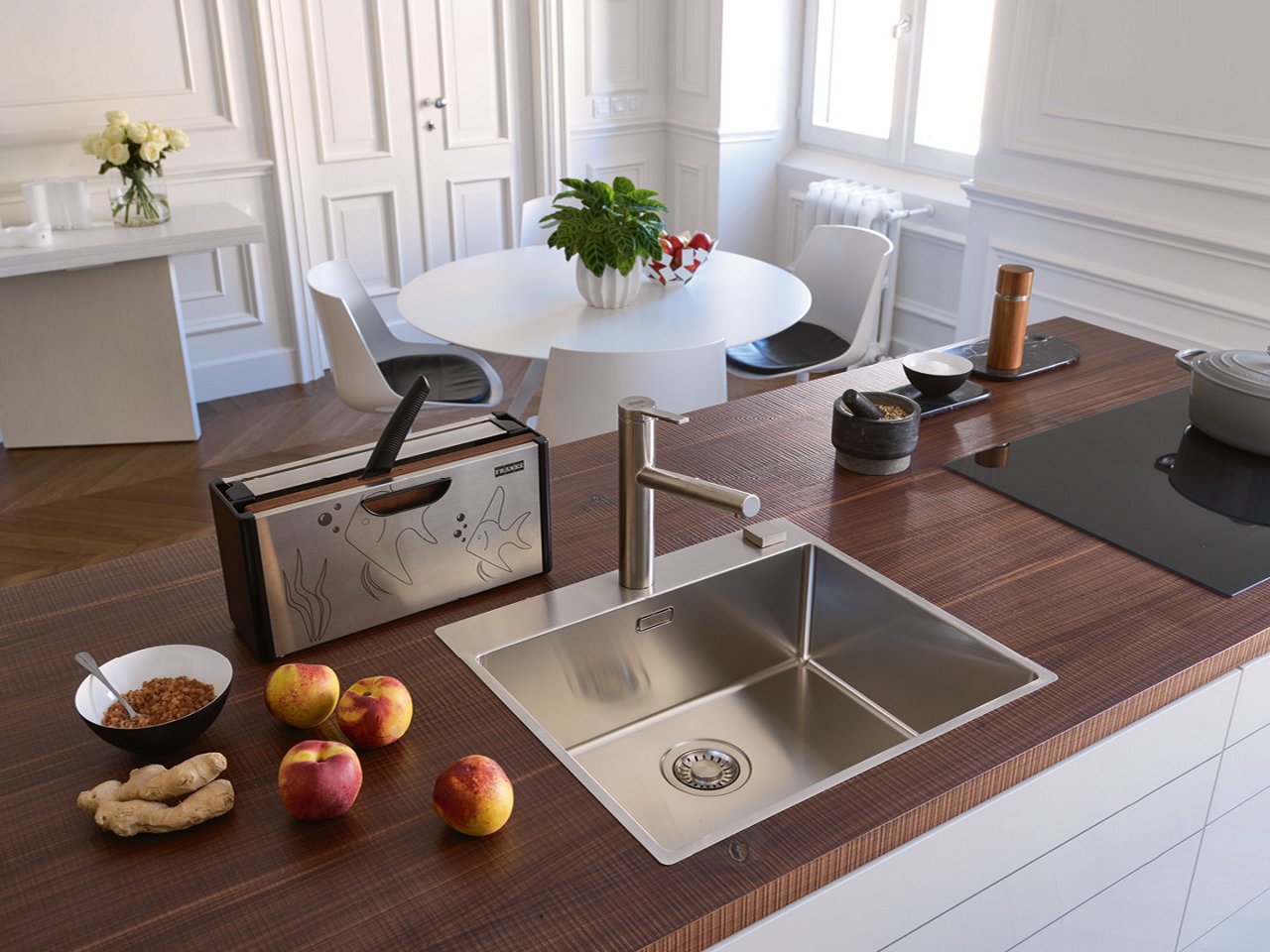Nowoczesna kuchnia z drewnianym blatem, na którym znajduje się stalowy zlewozmywak Franke z baterią, toster, trzy jabłka, korzeń imbiru oraz miska z płatkami; w tle widoczny biały stół z krzesłami i wazonem z kwiatami.
