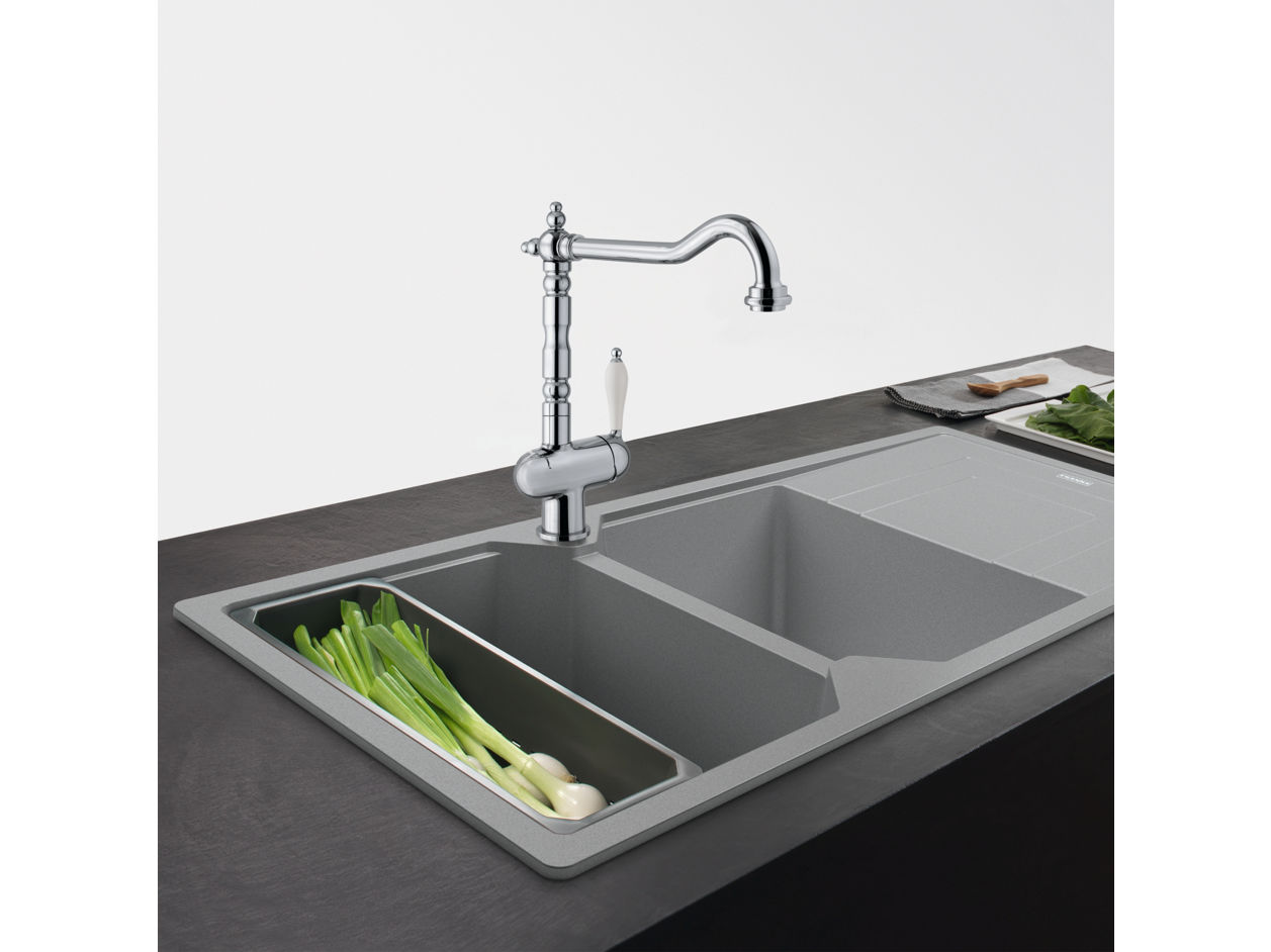 Nowoczesny zestaw zlewozmywakowy Franke z dwoma komorami – mniejsza z nich zawiera świeże zielone cebulki. Nad nią znajduje się chromowana bateria z białym uchwytem. W tle deska do krojenia z warzywami i nożem, na ciemnym blacie kuchennym.