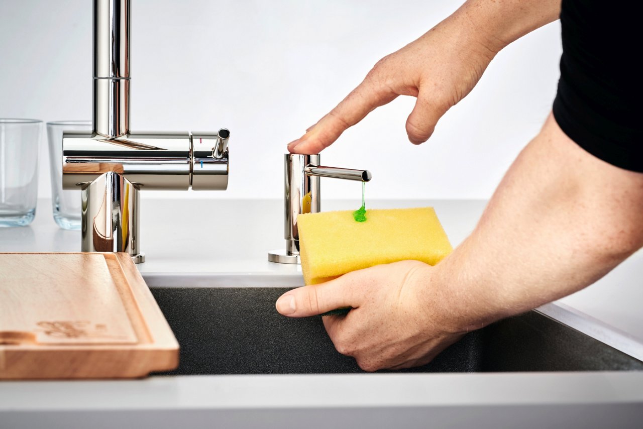 Zbliżenie na ręce osoby dozującej zielone mydło w płynie z chromowanego dozownika Franke na żółtą gąbkę nad zlewem kuchennym.