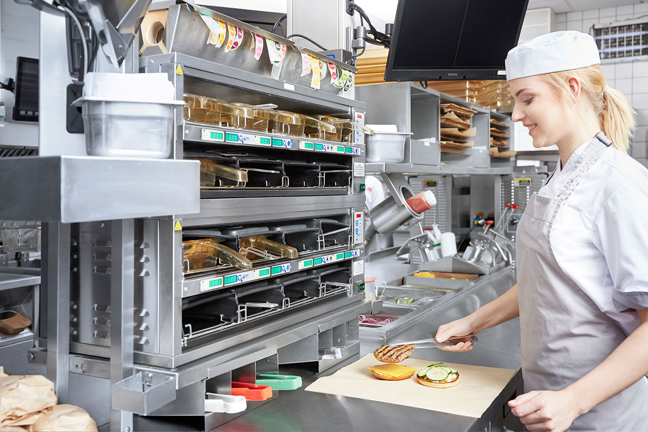 A person in a uniform and white cap assembles a sandwich in a fast food kitchen. They are placing vegetables on a bun in front of a large, stainless steel assembly station with trays and food storage compartments