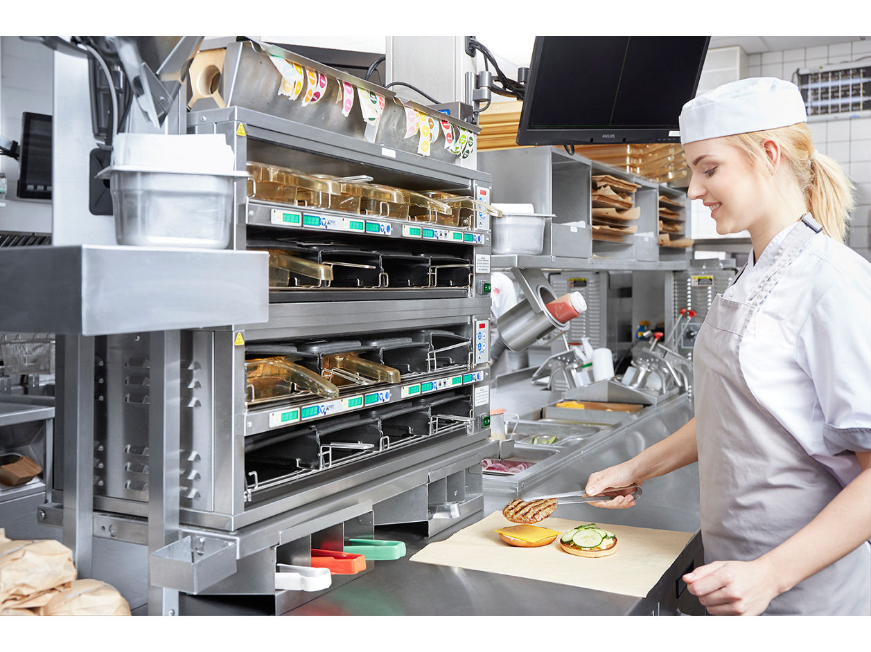 A person in a uniform and white cap assembles a sandwich in a fast food kitchen. They are placing vegetables on a bun in front of a large, stainless steel assembly station with trays and food storage compartments