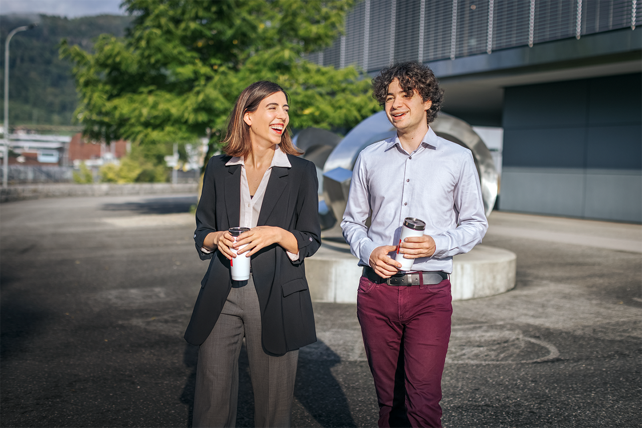 Two people walking outdoors, holding coffee cups, with a metallic sculpture and modern building in the background.