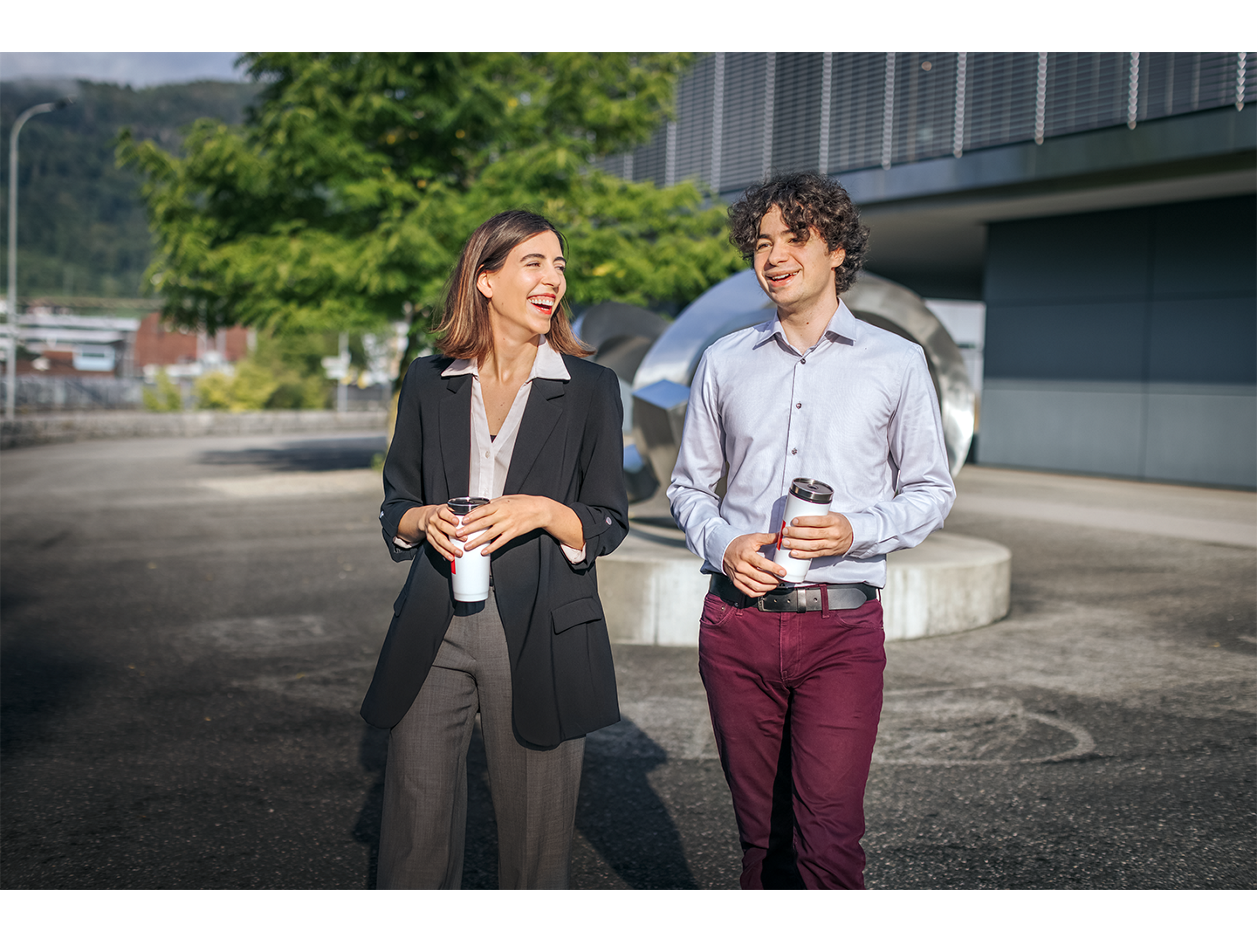 Two people walking outdoors, holding coffee cups, with a metallic sculpture and modern building in the background.