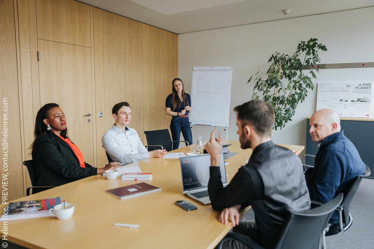 A meeting in a conference room with people seated around a table and a woman presenting at a flip chart.