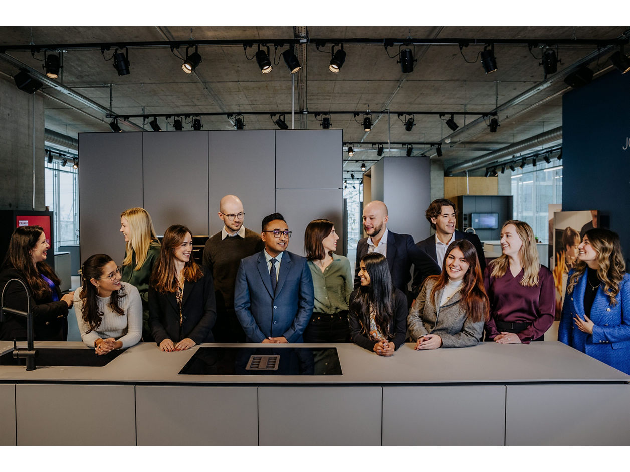 A group of fourteen people stand behind a Franke kitchen island in a modern, industrial-style room. They are dressed in business casual attire and appear engaged in conversation