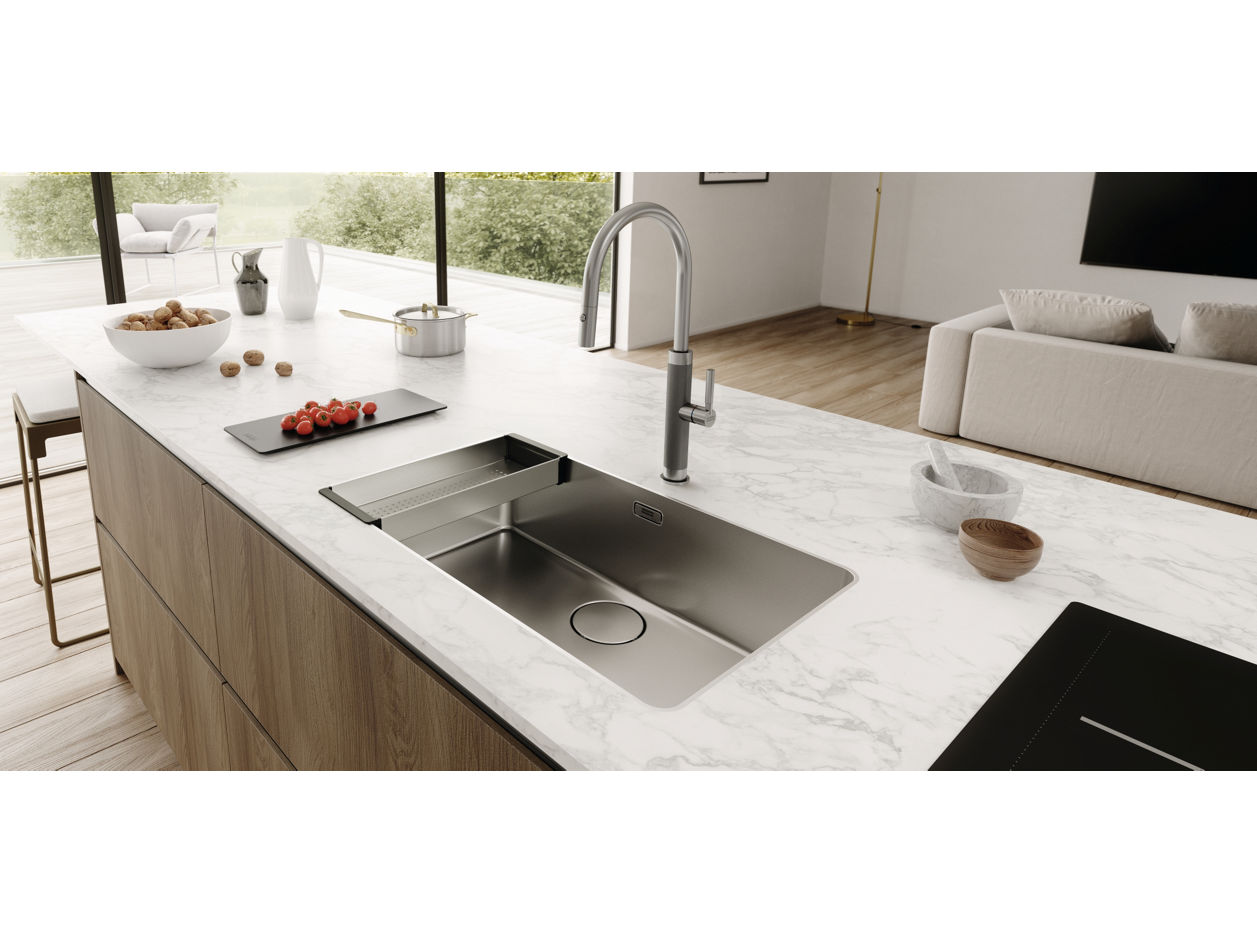 Nowoczesna kuchnia z marmurowym blatem, w którym osadzony jest elegancki, stalowy zlewozmywak i bateria Franke. Na blacie znajdują się m.in. miska z orzechami, deska do krojenia z truskawkami oraz mała drewniana miseczka. W tle widoczna biała sofa i duże okna z widokiem na zieleń.